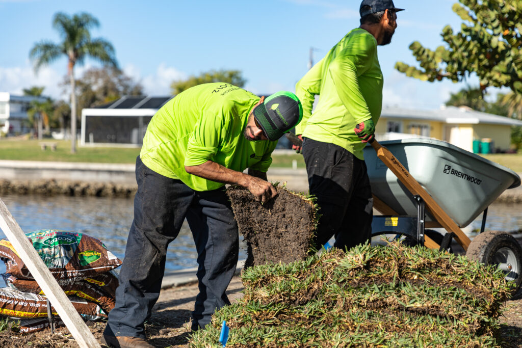 Two men in green shirts and yellow hats working on a pile of grass.